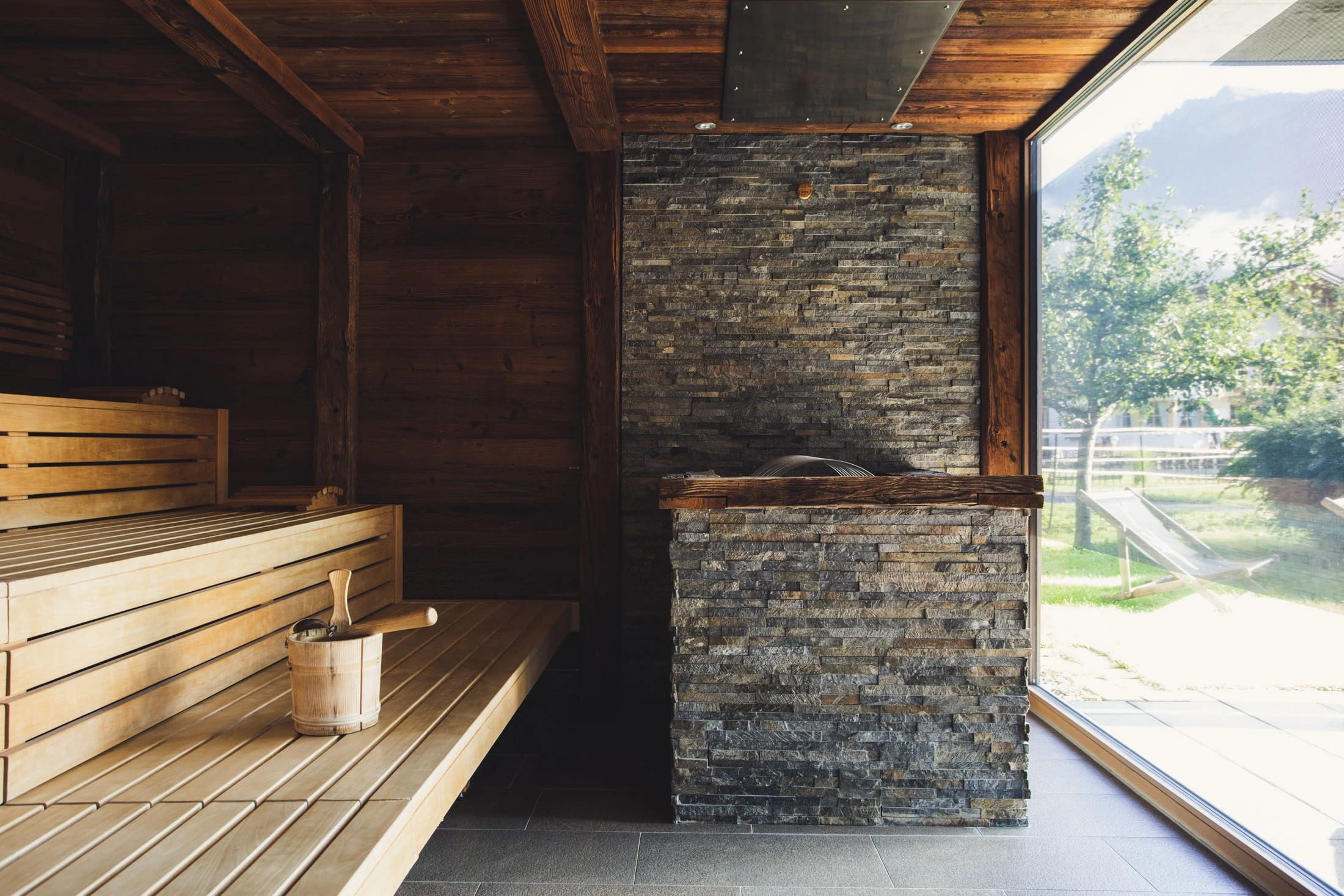 Sauna mit Holzbänken, Natursteinwand und großem Fenster ins Grüne