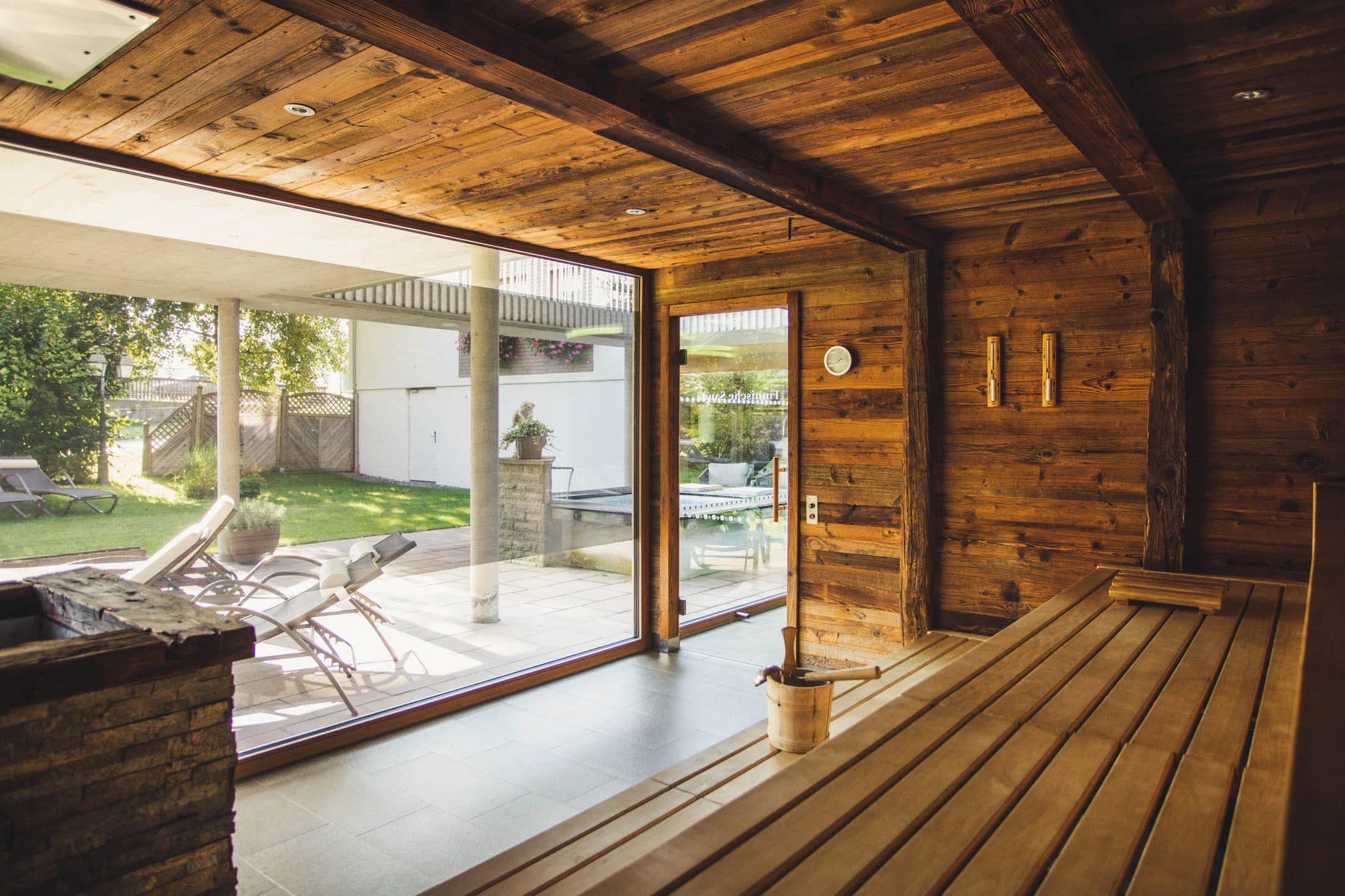 Sauna aus dunklem Holz mit geöffneter Tür zum Garten und Liegestühlen davor