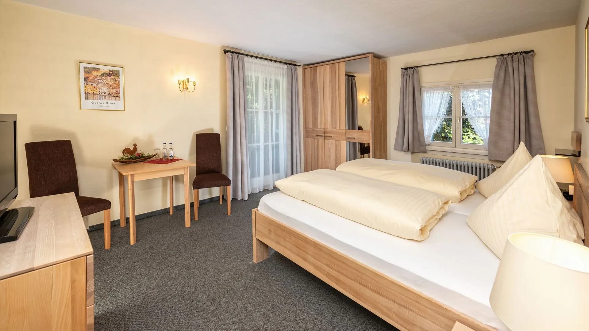 Doppelzimmer mit hellem Holzbett, Kleiderschrank, Fenster und kleiner Sitzecke