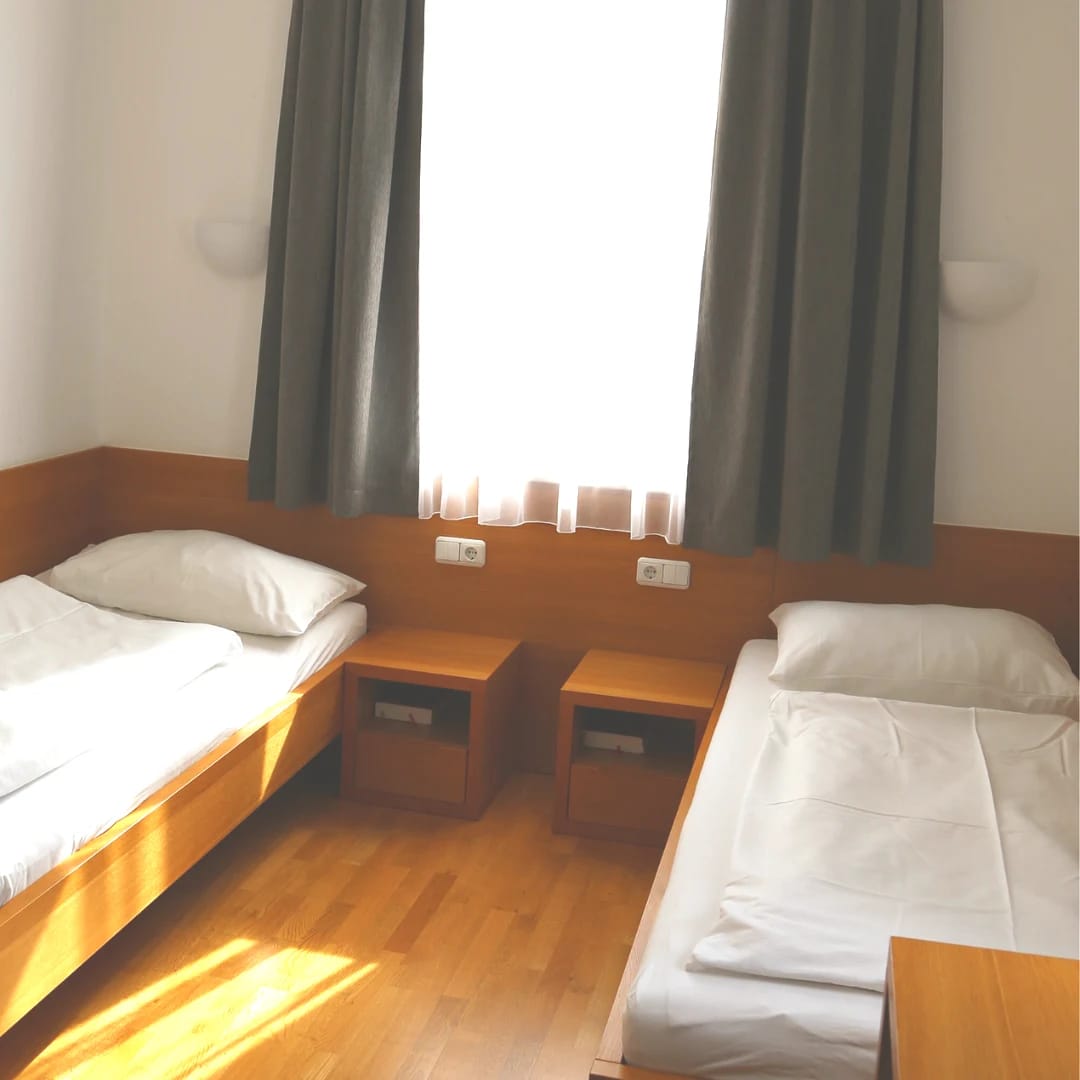Zimmer mit zwei Einzelbetten, Nachtkästchen, Holzboden und Fenster mit grauen Vorhängen