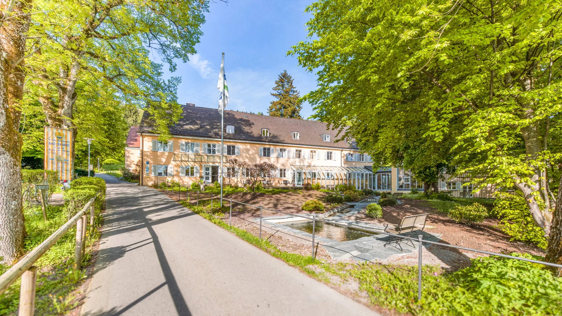 Zufahrtsweg zum Haupthaus mit heller Fassade, Fahnenmast und Wasserbecken zwischen hohen Bäumen