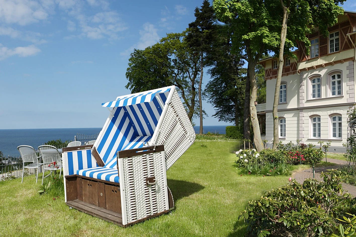 Blau-weißer Strandkorb auf der Rasenfläche im Garten, rechts das weiße Hotelgebäude, links Blick auf die Ostsee