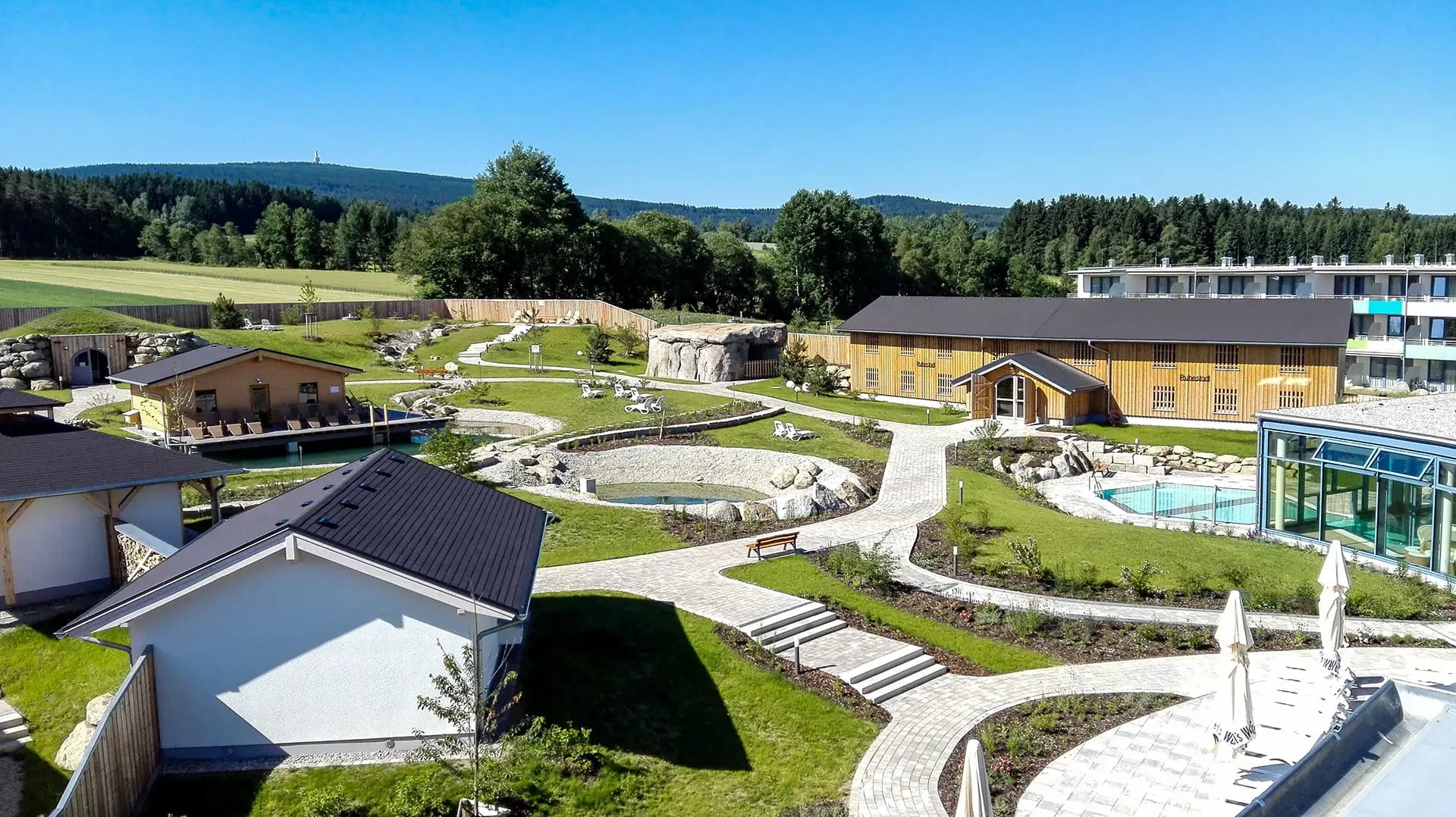 Saunagarten mit Holzhütten, angelegten Wegen und Blick auf das Hotelgebäude
