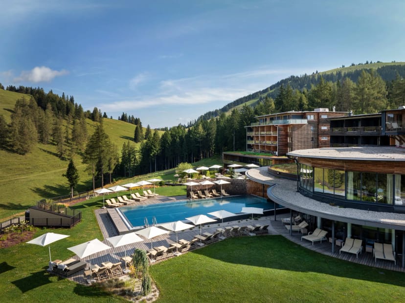 Hotelgebäude mit Außenpool, Sonnenliegen und Schirmen vor Almwiese und Nadelwald