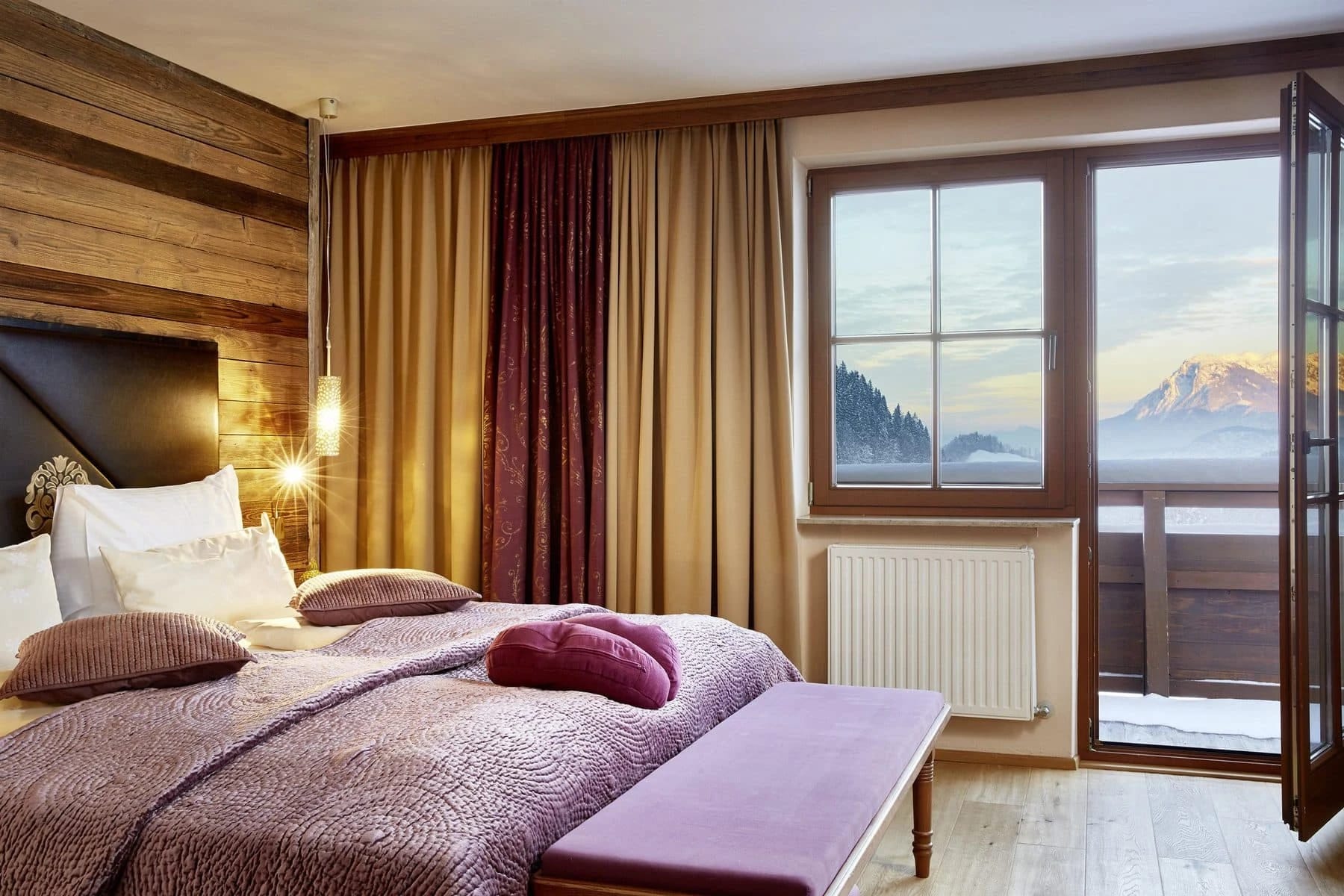Zimmer mit Holzwand, violetter Bettwäsche und Balkontür mit Blick auf verschneite Berge