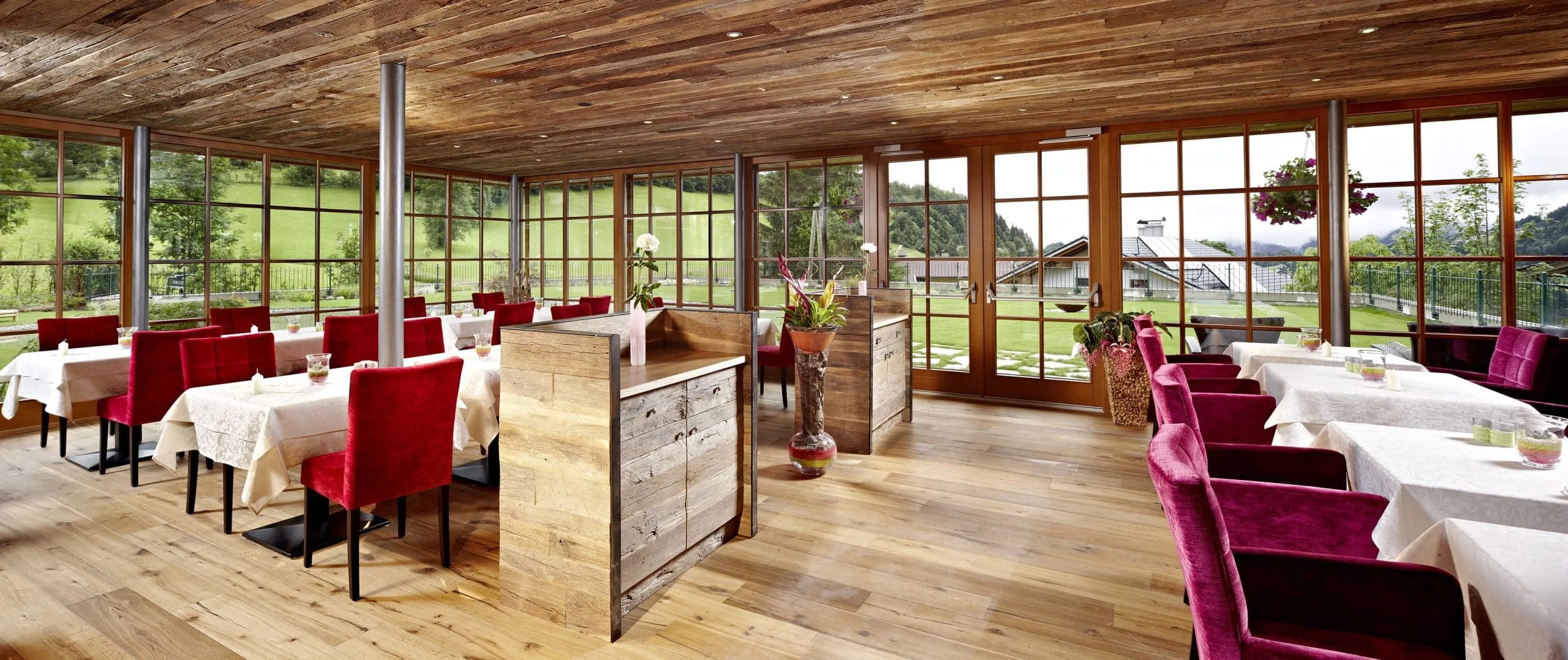 Wintergarten mit Holzdecke, eingedeckten Tischen, roten Stühlen und Panoramafenstern zum Garten