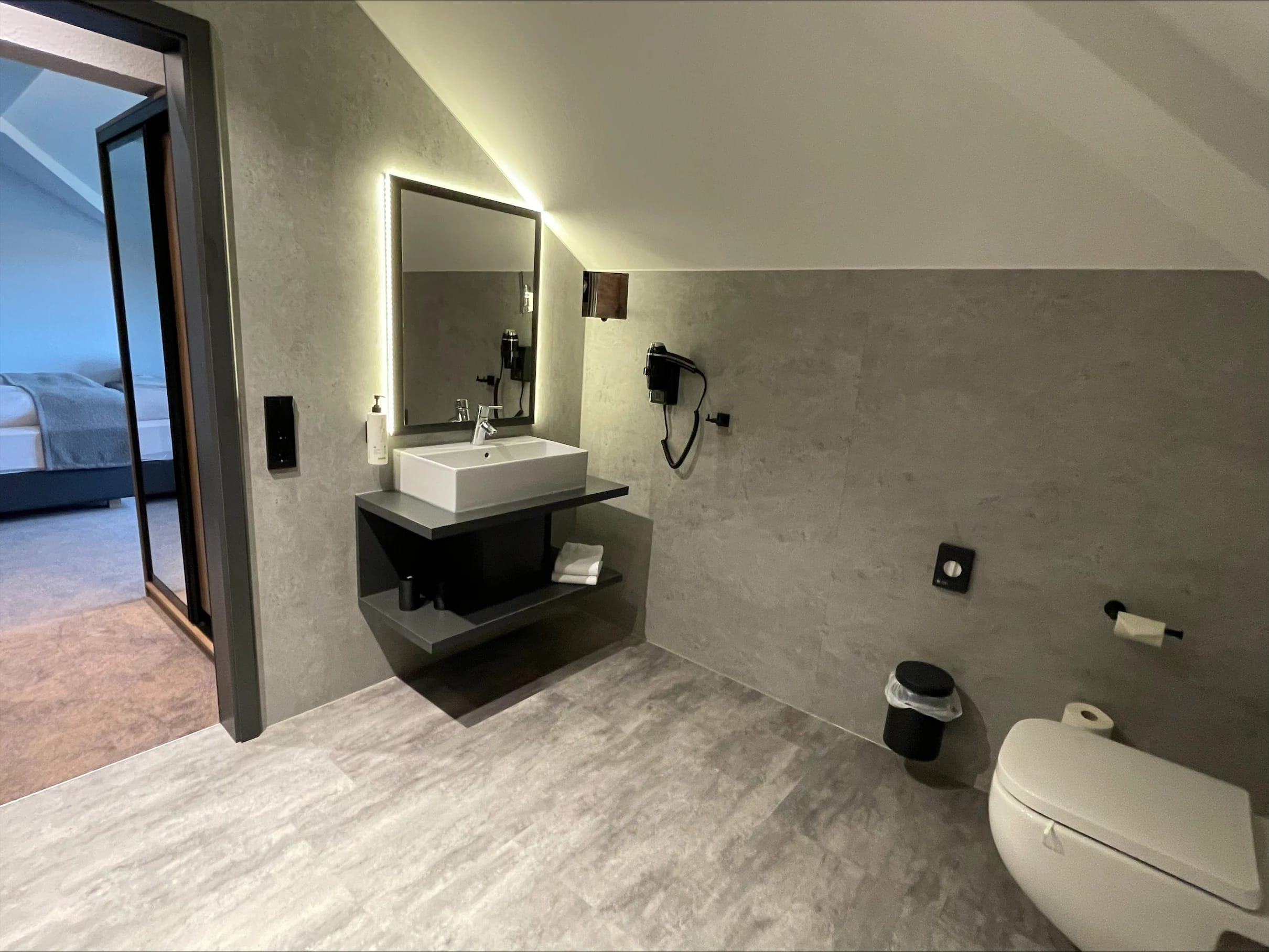 Modernes Badezimmer mit Waschtisch, beleuchtetem Spiegel und WC, durch die offene Tür der Blick ins Zimmer