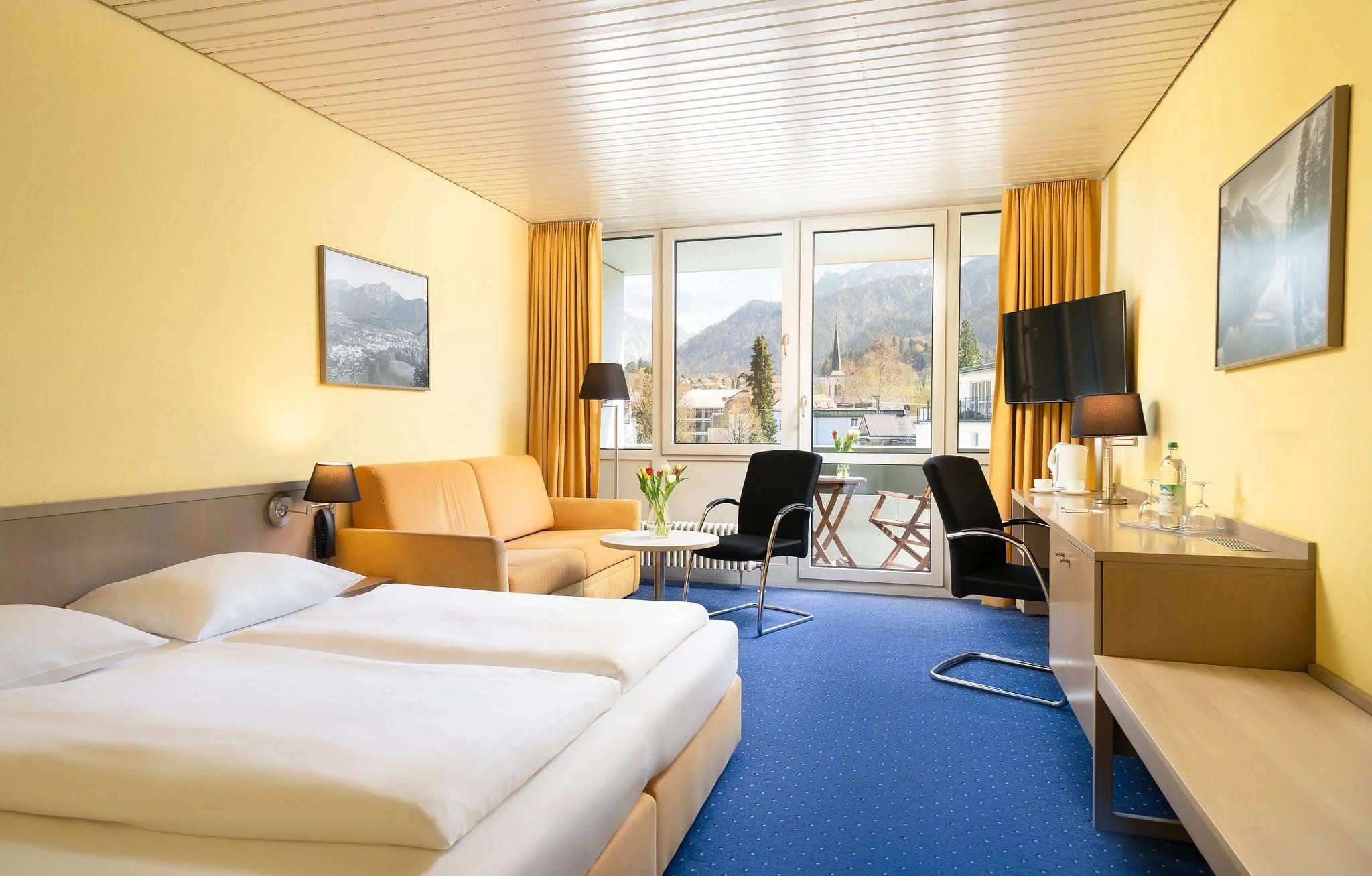 Helles Zimmer mit Doppelbett, Sofa und Fensterfront mit Bergblick