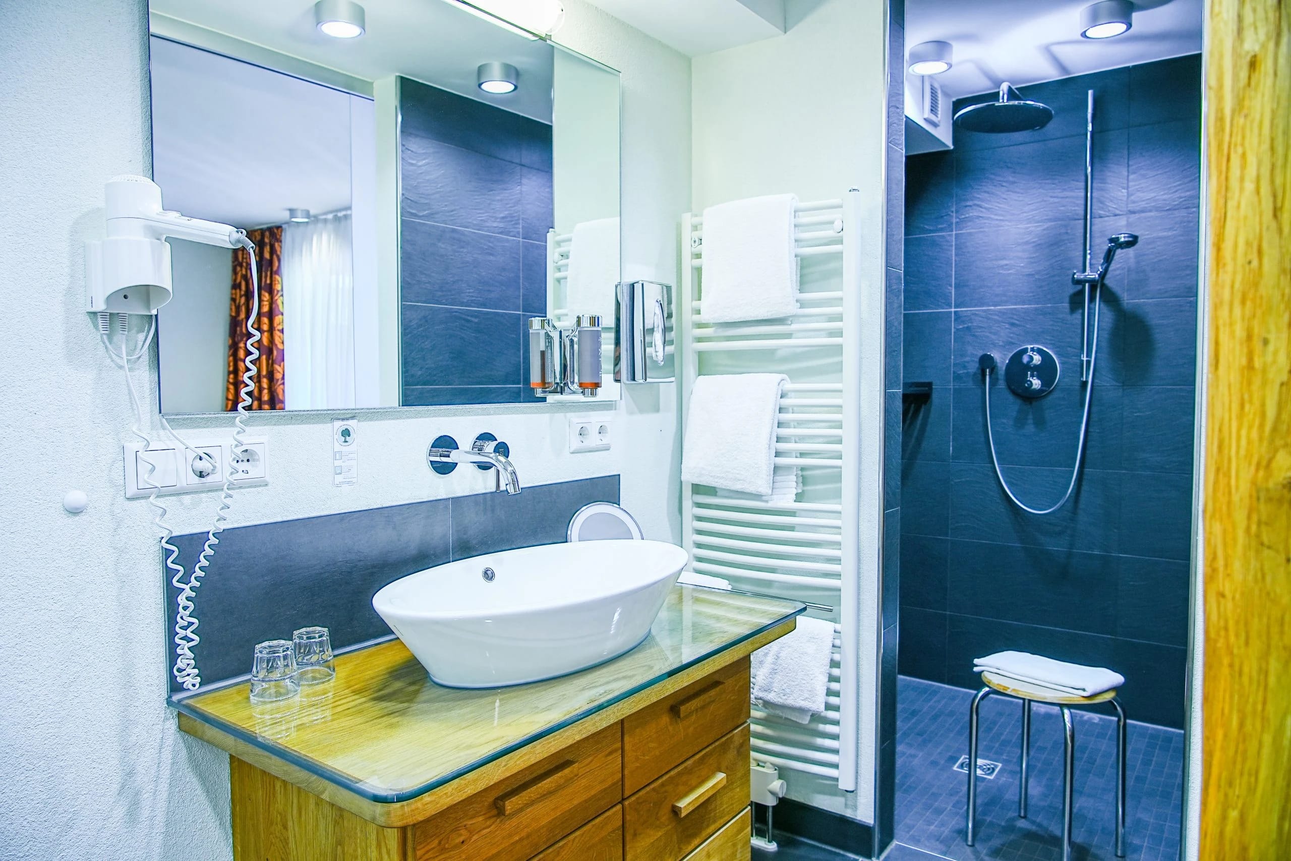Badezimmer mit Aufsatzwaschbecken auf Holzkonsole, großem Spiegel und bodengleicher Dusche mit dunklen Fliesen