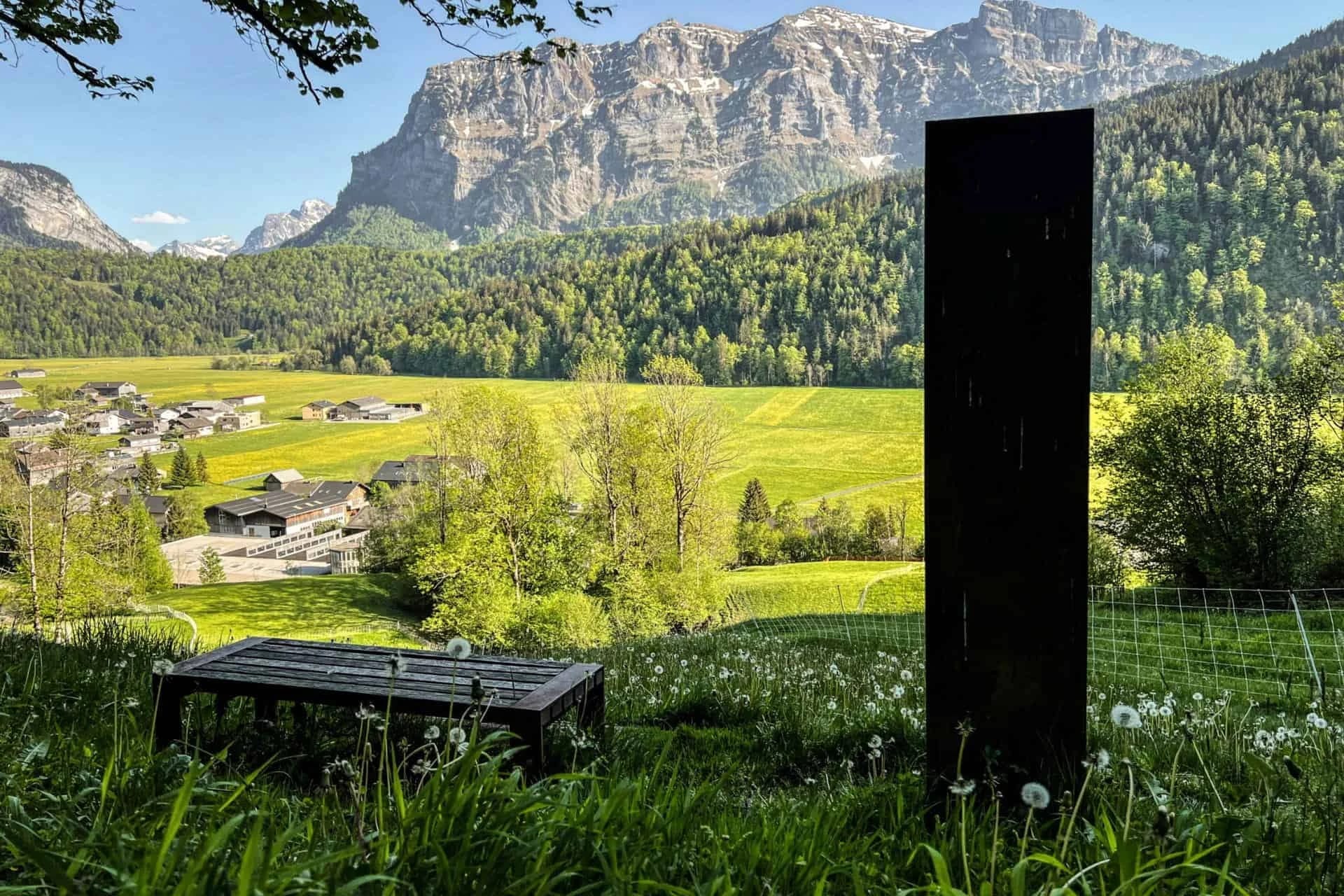 Blick über grüne Wiesen und das Dorf auf eine hohe Felswand, im Vordergrund eine Bank