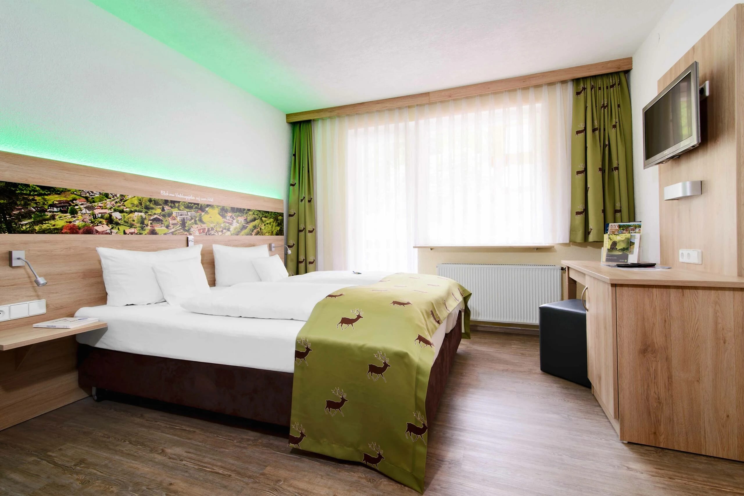 Modernes Doppelzimmer mit grünen Vorhängen, indirekter Beleuchtung und Waldmotiv über dem Bett