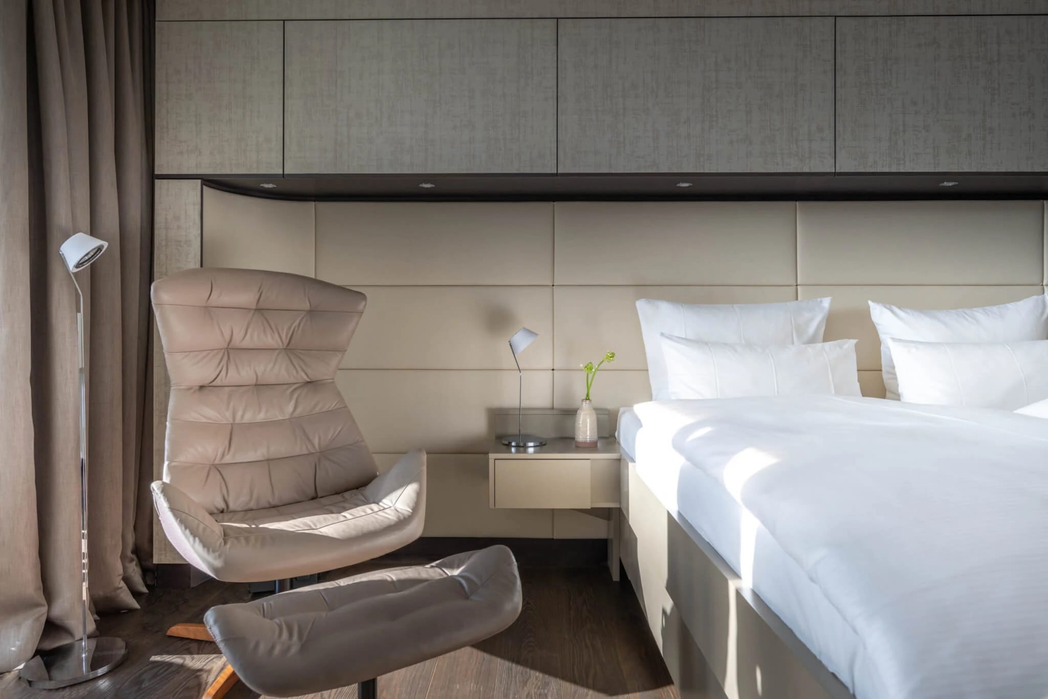 Schlafzimmer mit Doppelbett, gepolstertem Kopfteil, Nachttisch und Loungesessel mit Hocker