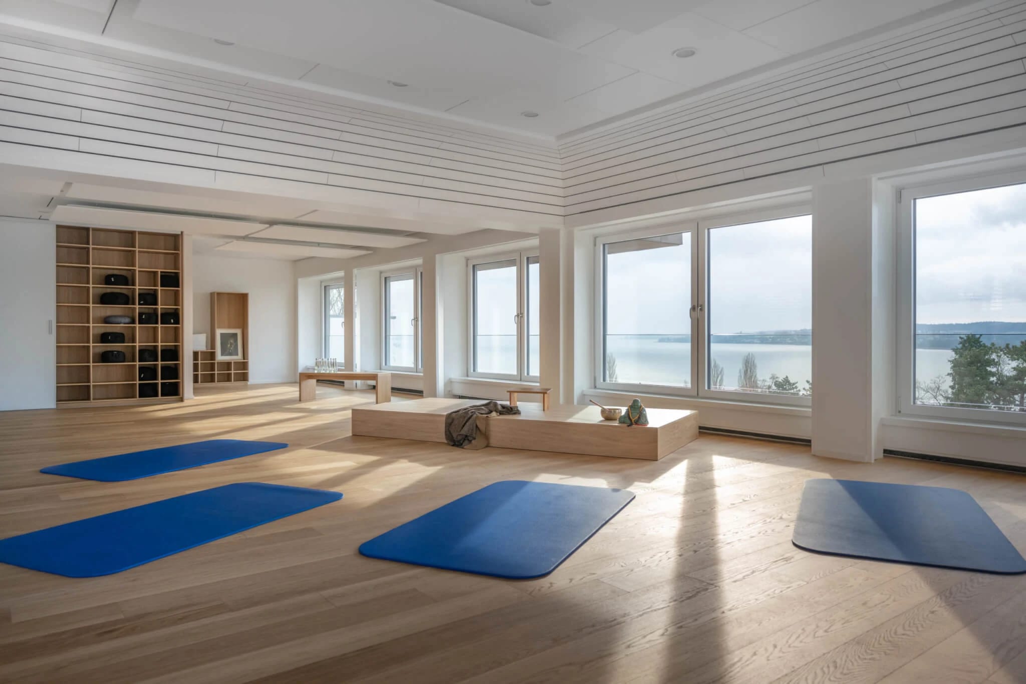 Heller Gymnastikraum mit Holzboden, ausgelegten Yogamatten und Fensterfront zum See