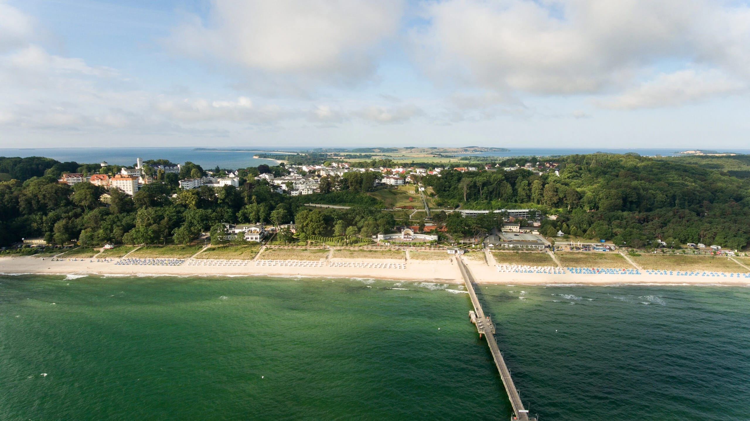 Luftbild der Küste mit langem Sandstrand, Strandkorbreihen, Seebrücke und bewaldetem Steilhang