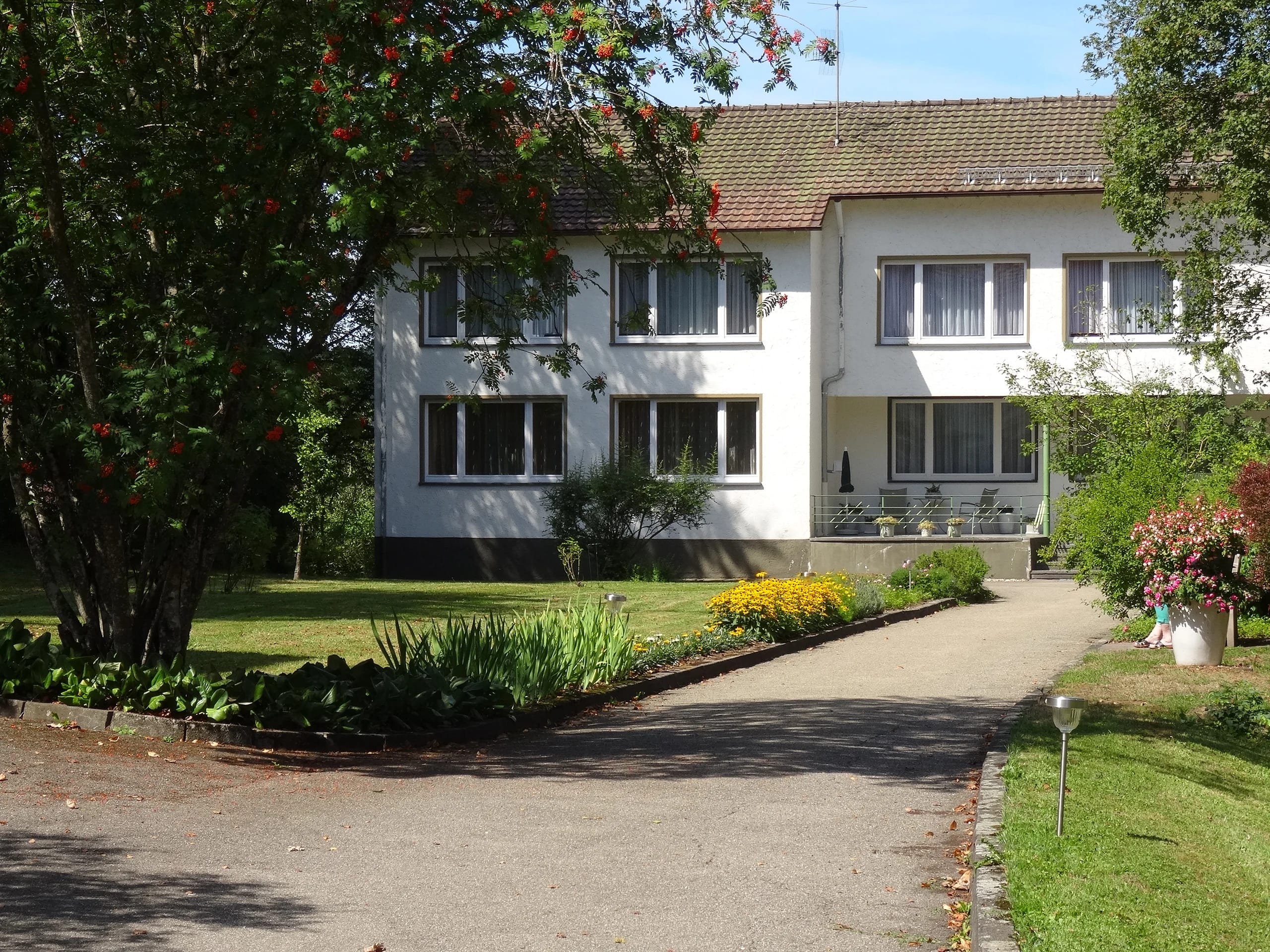 Weißes Haus mit dunklem Dach, davor Zufahrt, Blumenbeet und Rasen bei Sonnenschein