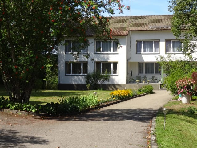 Weißes Haus mit dunklem Dach, davor Zufahrt, Blumenbeet und Rasen bei Sonnenschein