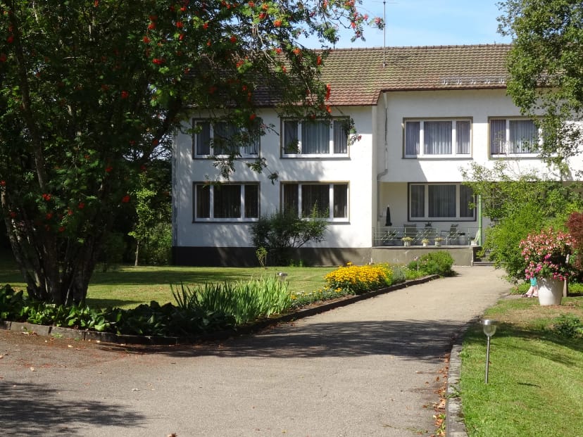 Weißes Haus mit dunklem Dach, davor Zufahrt, Blumenbeet und Rasen bei Sonnenschein