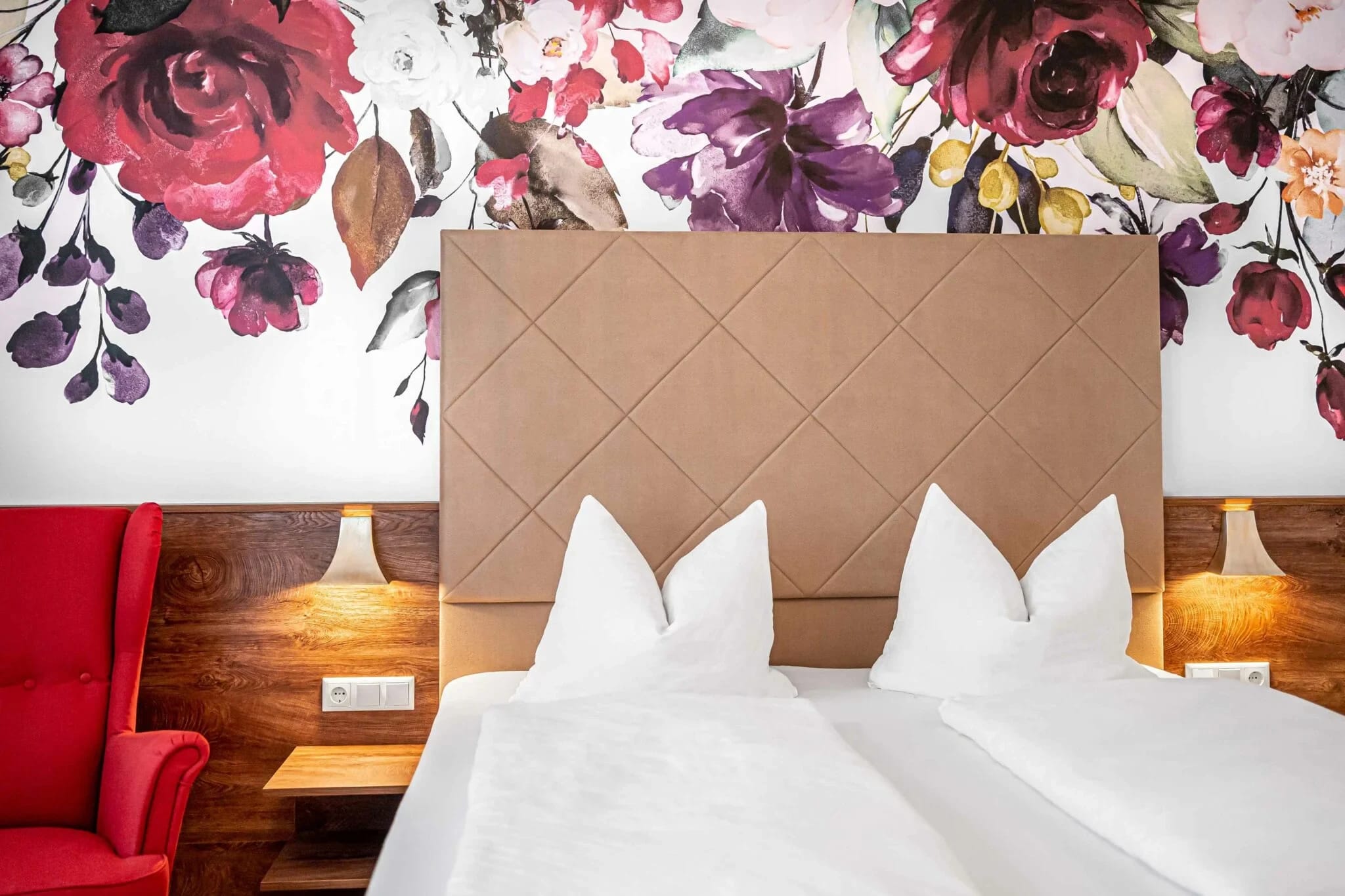 Doppelbett mit gepolstertem Kopfteil vor floraler Wandtapete, daneben ein roter Sessel