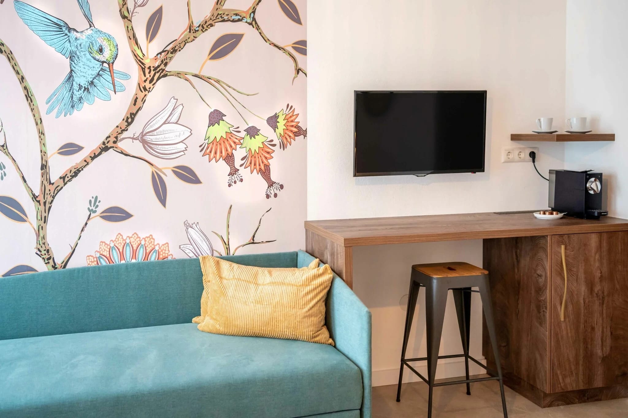 Zimmer mit Vogelmotiv-Tapete, türkisem Sofa, Holzschreibtisch und Fernseher