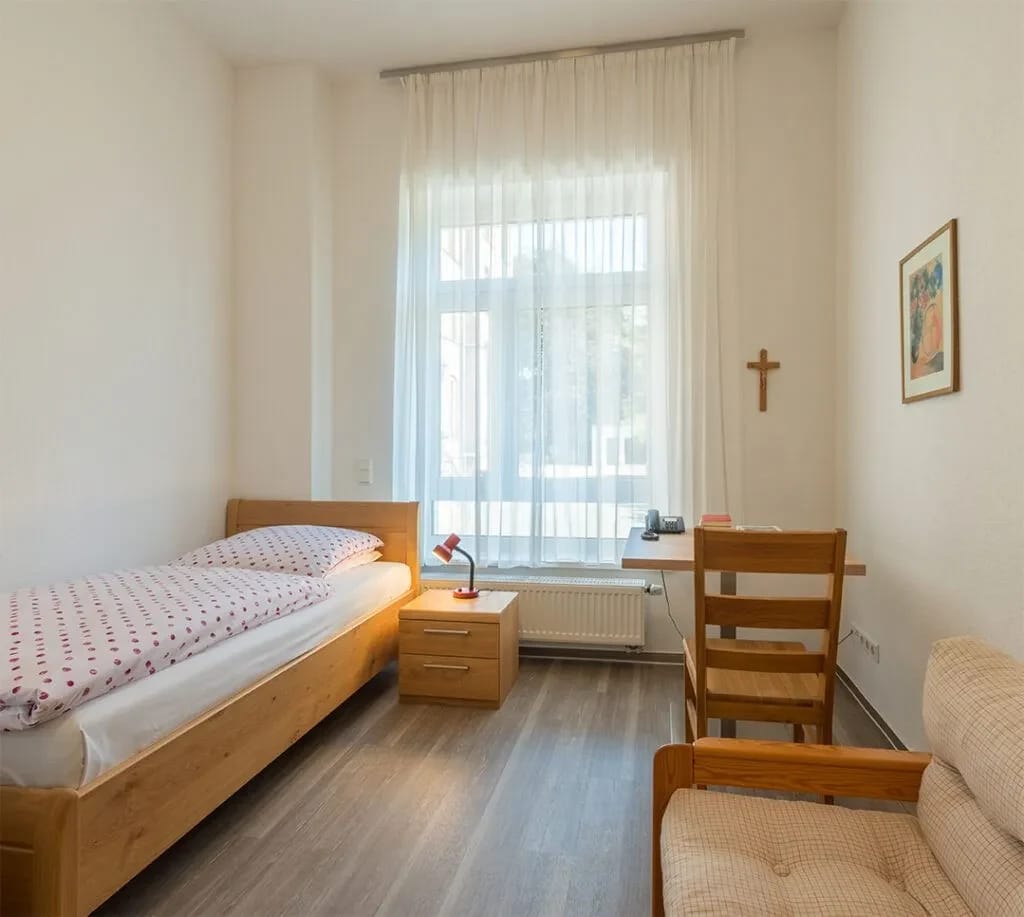 Einzelzimmer mit Bett, Nachttisch und Kruzifix an der Wand