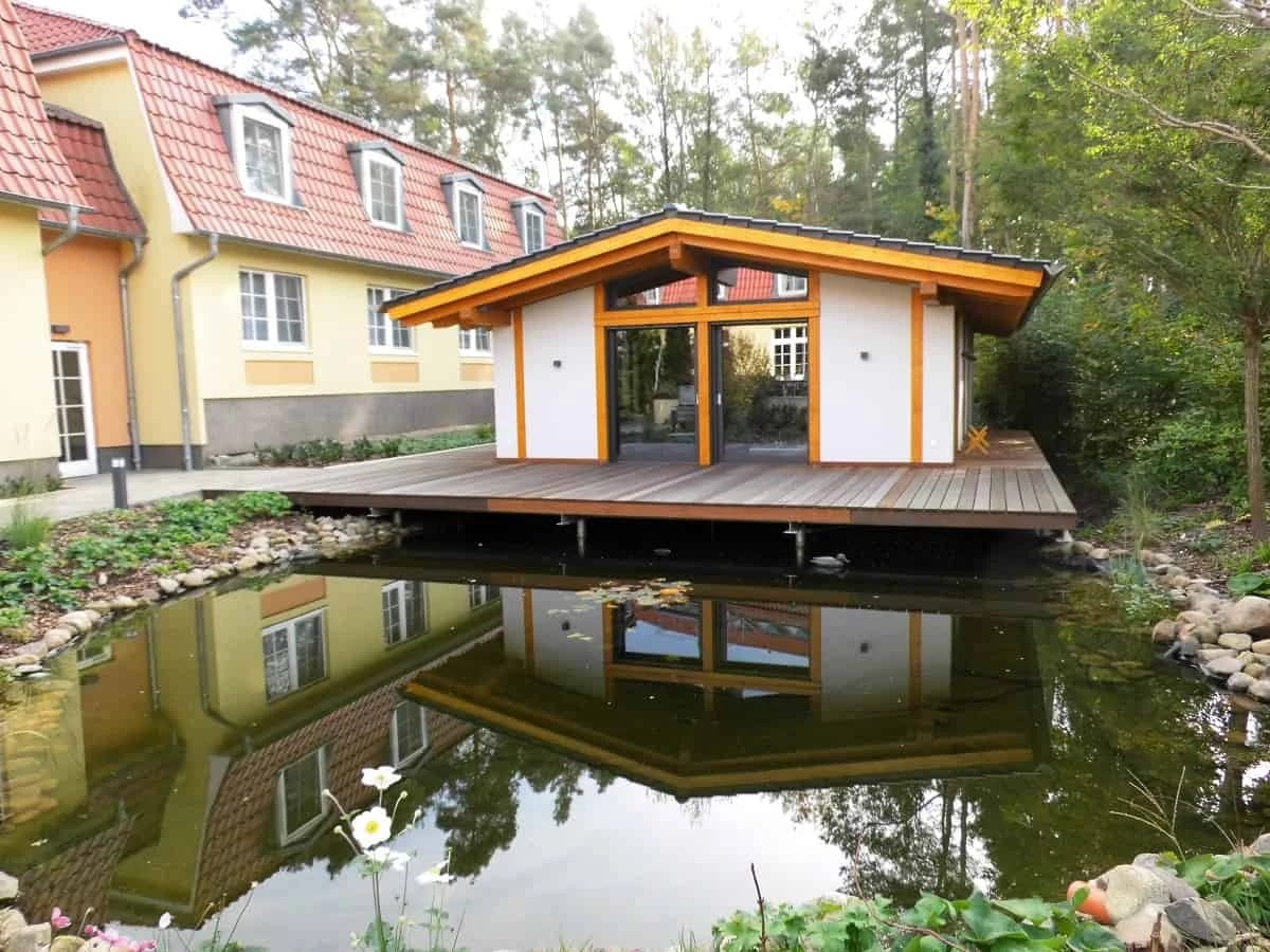 Saunahaus mit Holzterrasse am Teich, dahinter das Hotelgebäude