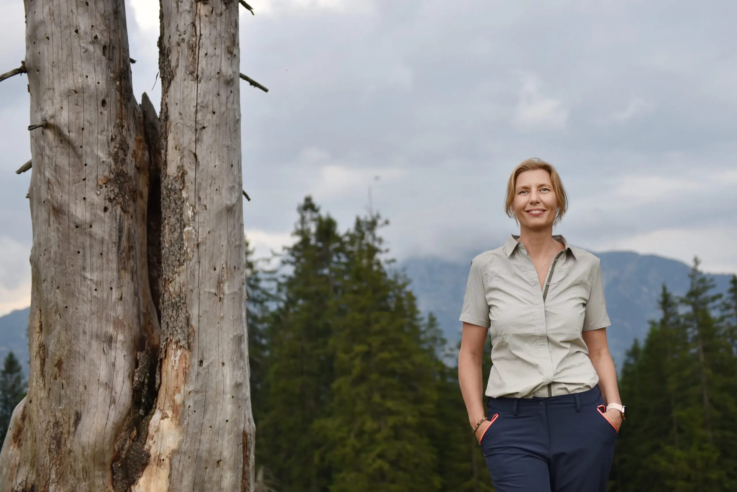 Frau in Outdoorkleidung steht vor Bergpanorama neben einem abgestorbenen Baum