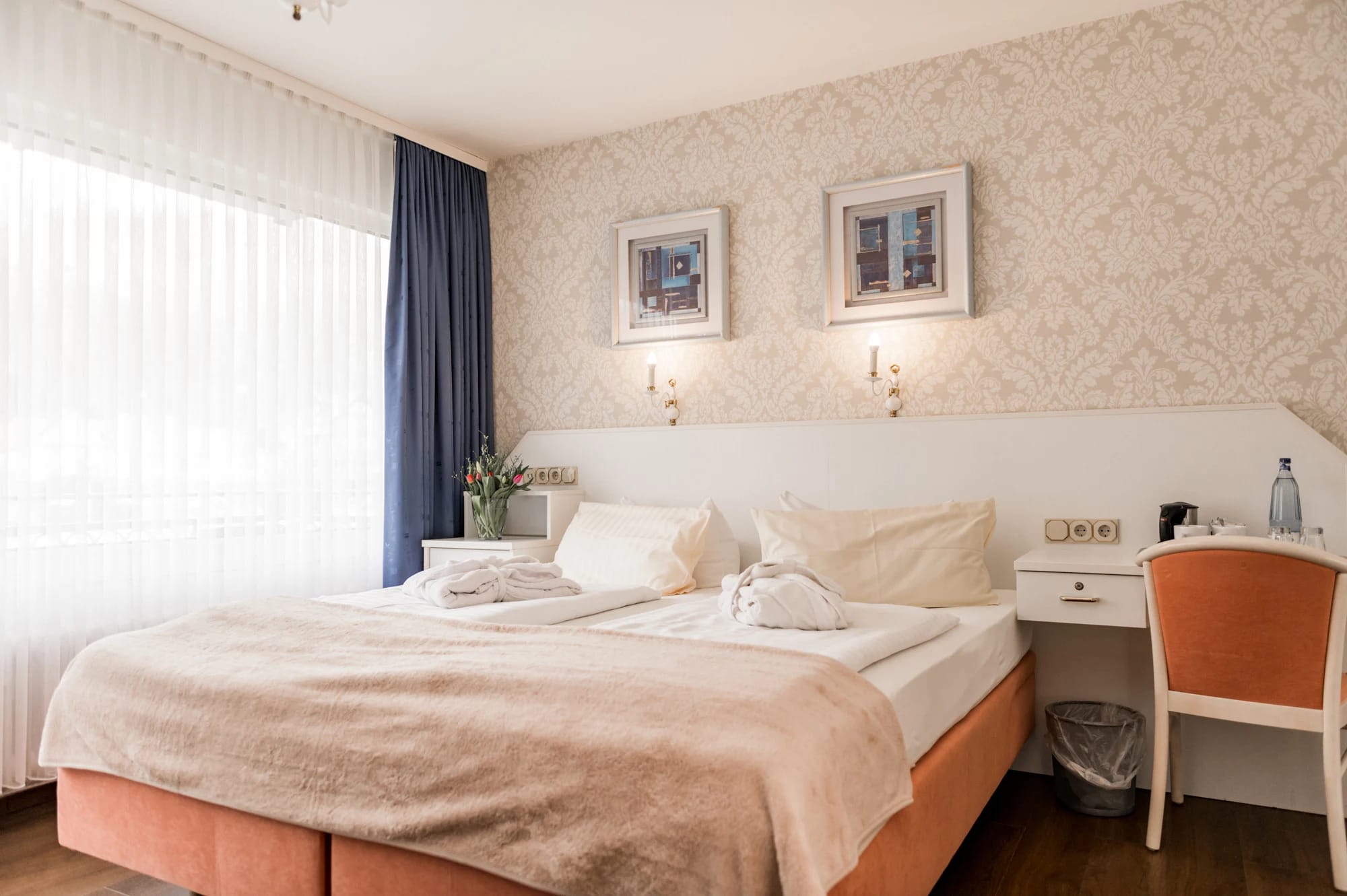 Doppelzimmer mit gemusterter Tapete, zwei Bildern über dem Bett und blauem Vorhang