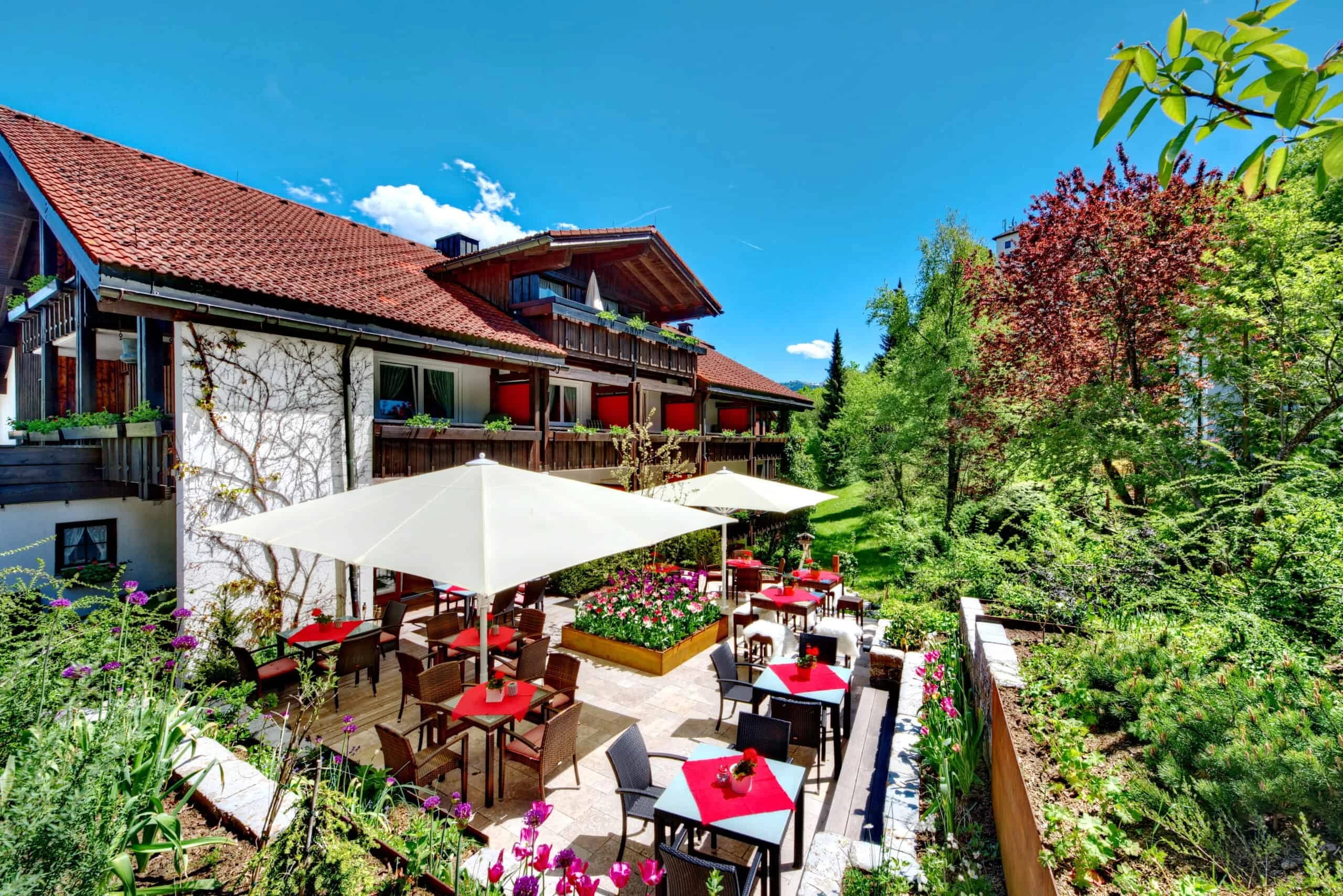Terrasse mit roten Tischen und Sonnenschirmen vor dem Haus, umgeben von Blumenbeeten