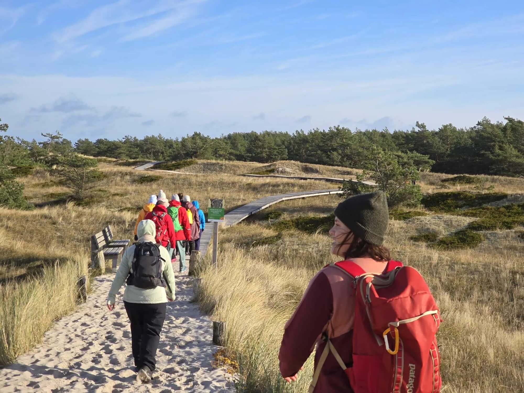 Fastenwandergruppe mit bunten Rucksäcken auf einem Bohlenweg durch die Dünen