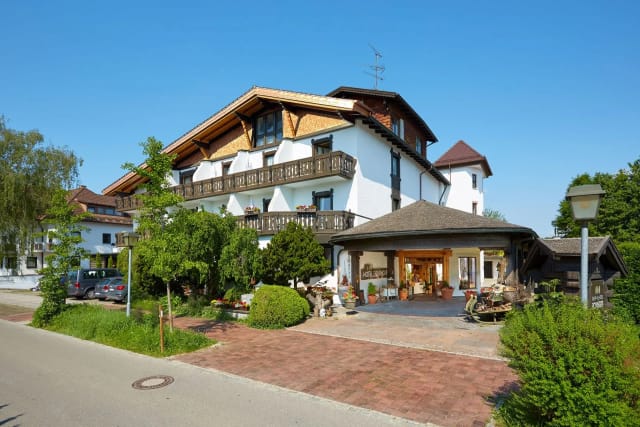 Weißes Hotelgebäude mit Holzbalkonen, überdachter Eingang und gepflasterter Vorfahrt bei Sonnenschein