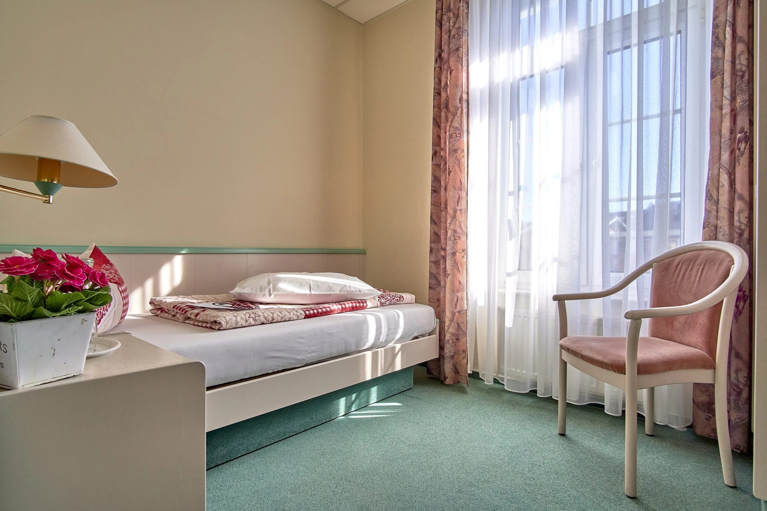 Einzelzimmer mit schmalem Bett, Sessel und sonnenbeschienenem Fenster