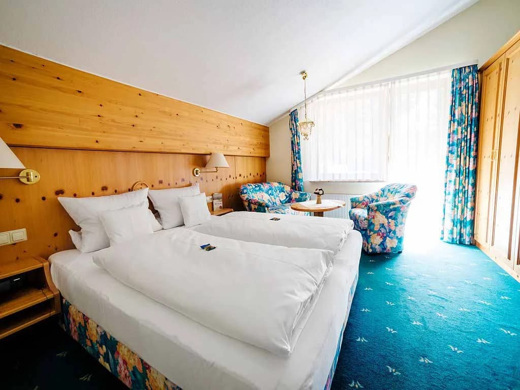Schlafzimmer mit Holzvertäfelung, zwei zusammengestellten Betten, Sesseln am Fenster und türkisfarbenem Teppichboden