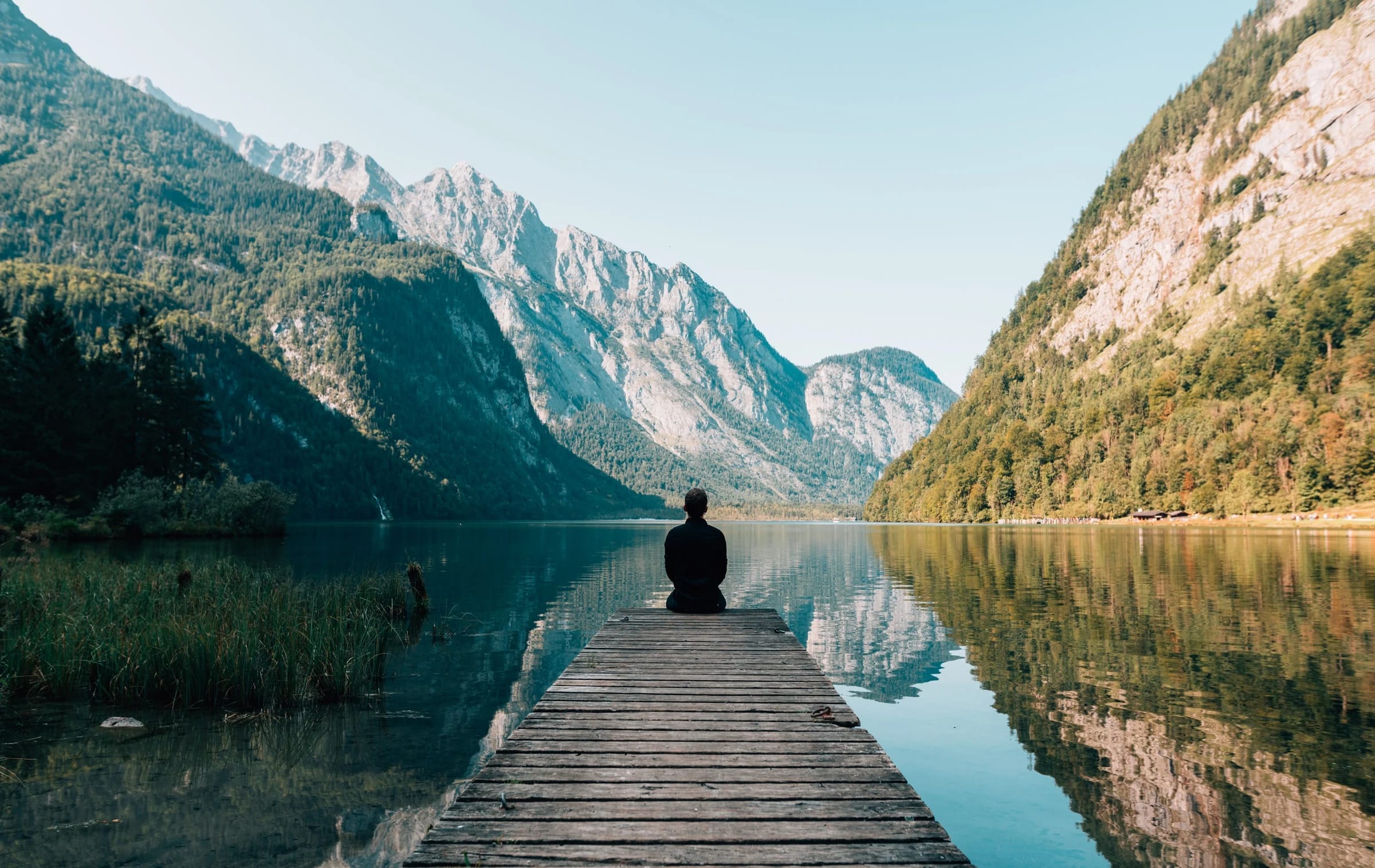 Person sitzt meditierend auf einem Holzsteg an einem Bergsee, umgeben von Bergen