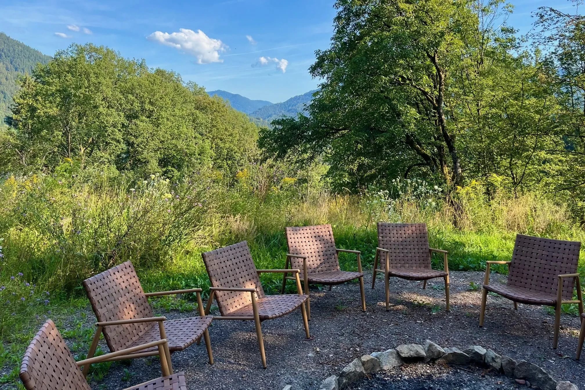 Feuerstelle im Garten mit Sesseln im Kreis, dahinter Wiese, Bäume und Blick auf die Berge