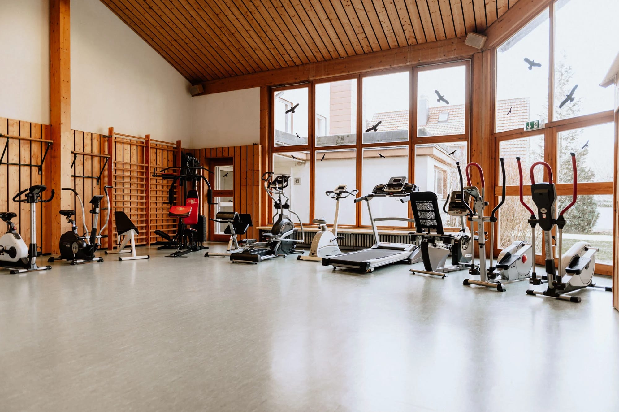 Fitnessraum mit Ausdauergeräten unter Holzdecke und hohen Fenstern