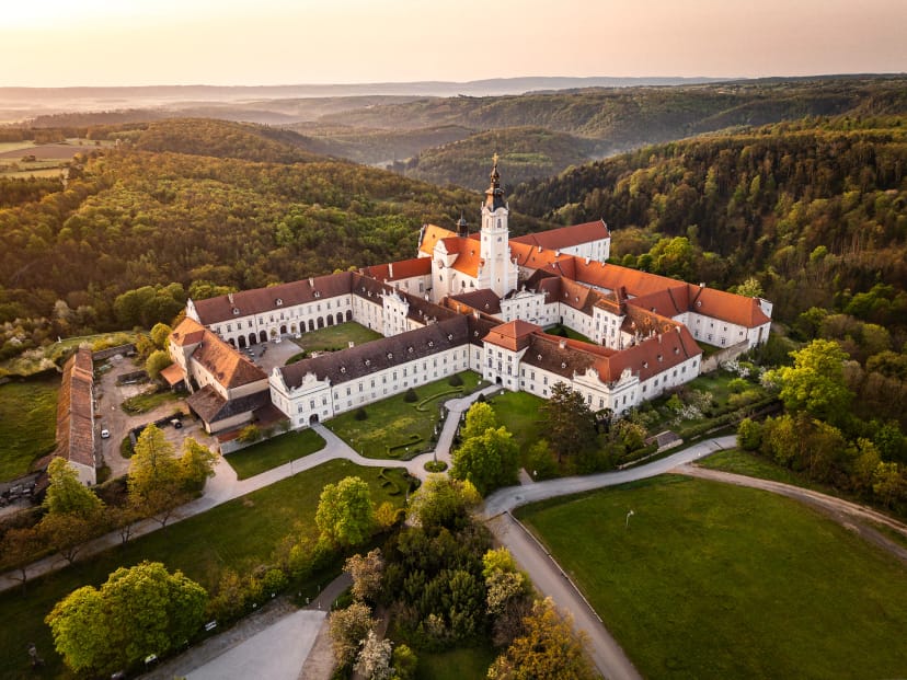Luftaufnahme der barocken Stiftsanlage mit roten Ziegeldächern und Kirchturm, umgeben von bewaldeten Hügeln im Abendlicht