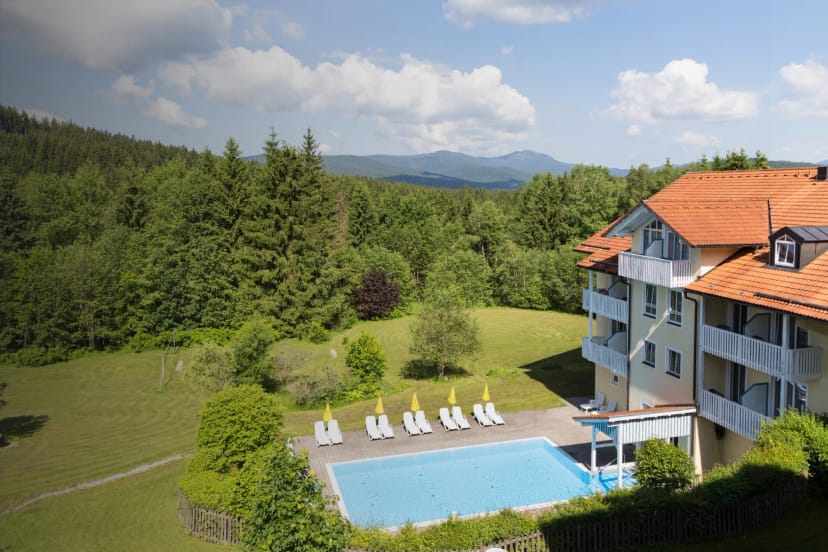 Luftaufnahme des Hotels mit Außenpool, Liegewiese und bewaldeten Bergen im Hintergrund