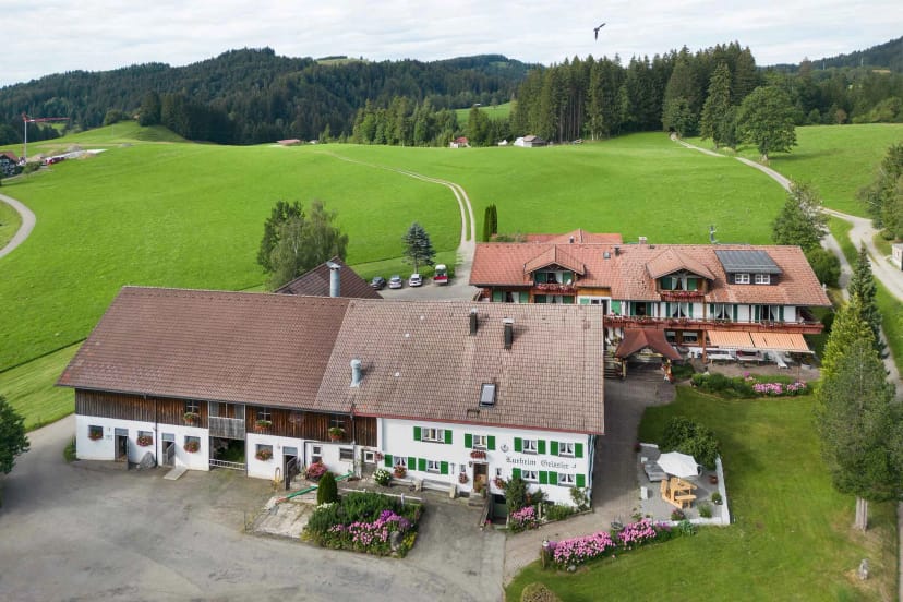 Luftaufnahme des Hofes mit langgestrecktem weißen Bauernhaus und angebautem Gästehaus inmitten grüner Wiesen