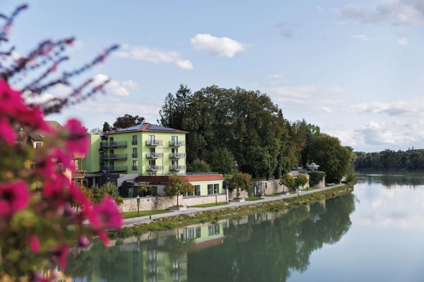 Grün gestrichenes Hotelgebäude direkt am Ufer des Inn, im Vordergrund blühende Sträucher