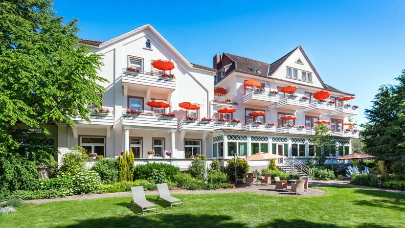 Weiße Hotelfassade mit Balkonen und orangefarbenen Sonnenschirmen, davor eine Rasenfläche mit zwei Sonnenliegen
