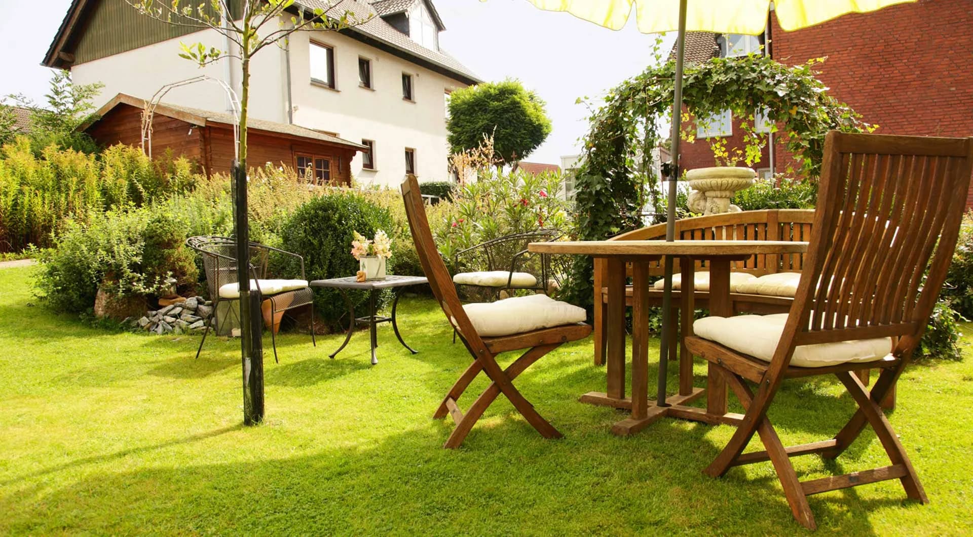 Garten mit Rasen, Holztisch und Gartenstühlen, dahinter Sträucher und Häuser