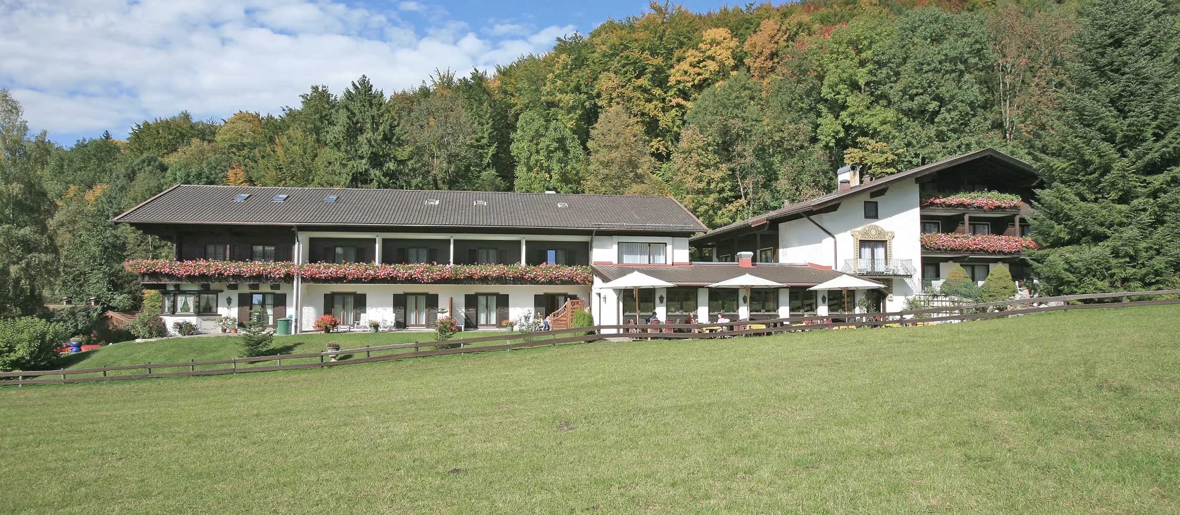 Gesamtansicht des Hauses mit Terrasse und Sonnenschirmen, davor eine große Wiese, dahinter ein bewaldeter Hang