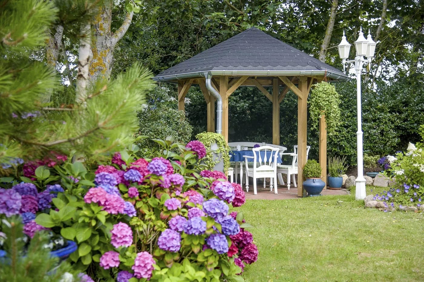 Überdachter Holzpavillon mit Sitzgruppe im Garten, umgeben von blühenden Hortensien