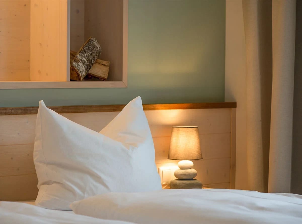 Bettdetail mit weißem Kissen, Nachttischlampe und Holzregal mit Feuerholz