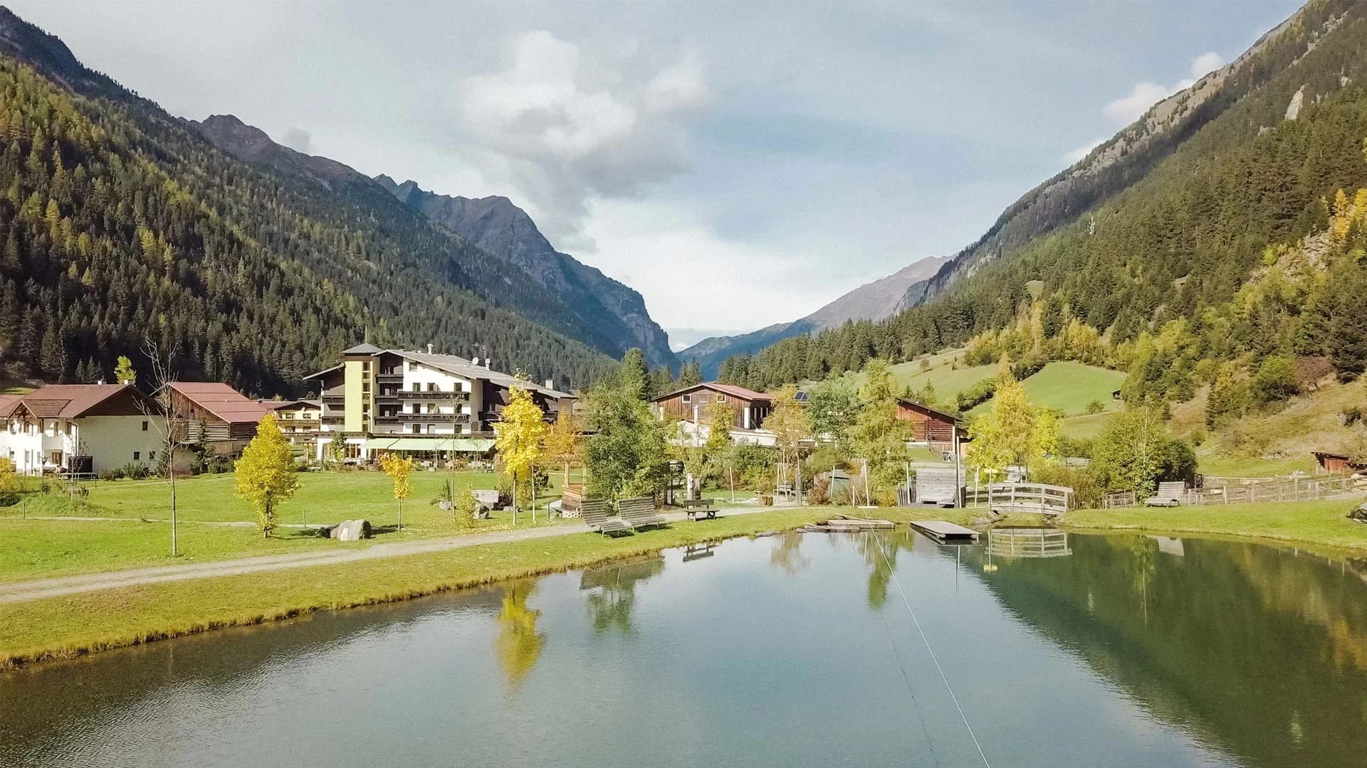 Das Hotel im Tal mit kleinem See im Vordergrund und bewaldeten Berghängen