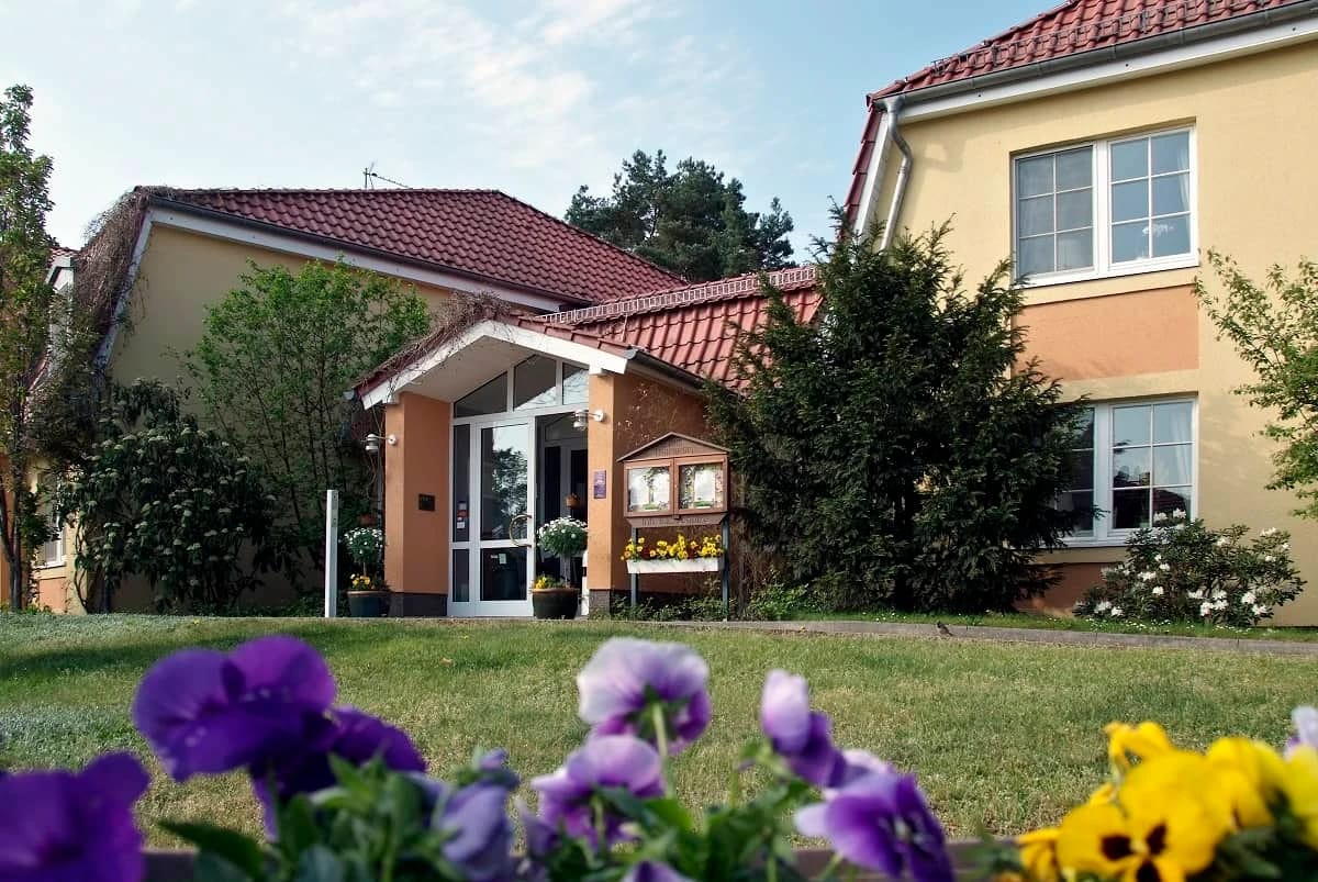 Außenansicht des Hotels mit überdachtem Eingang, Rasenfläche und blühenden Stiefmütterchen