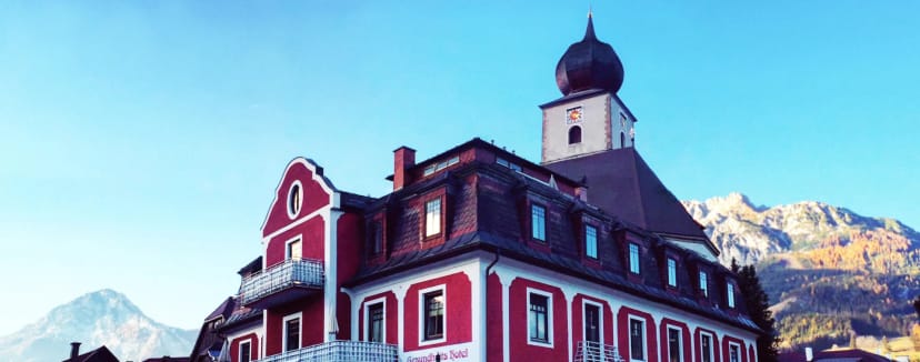 Rot-weiße Fassade des Hauses mit Balkon, dahinter ein Kirchturm mit Zwiebelhaube und verschneite Berge