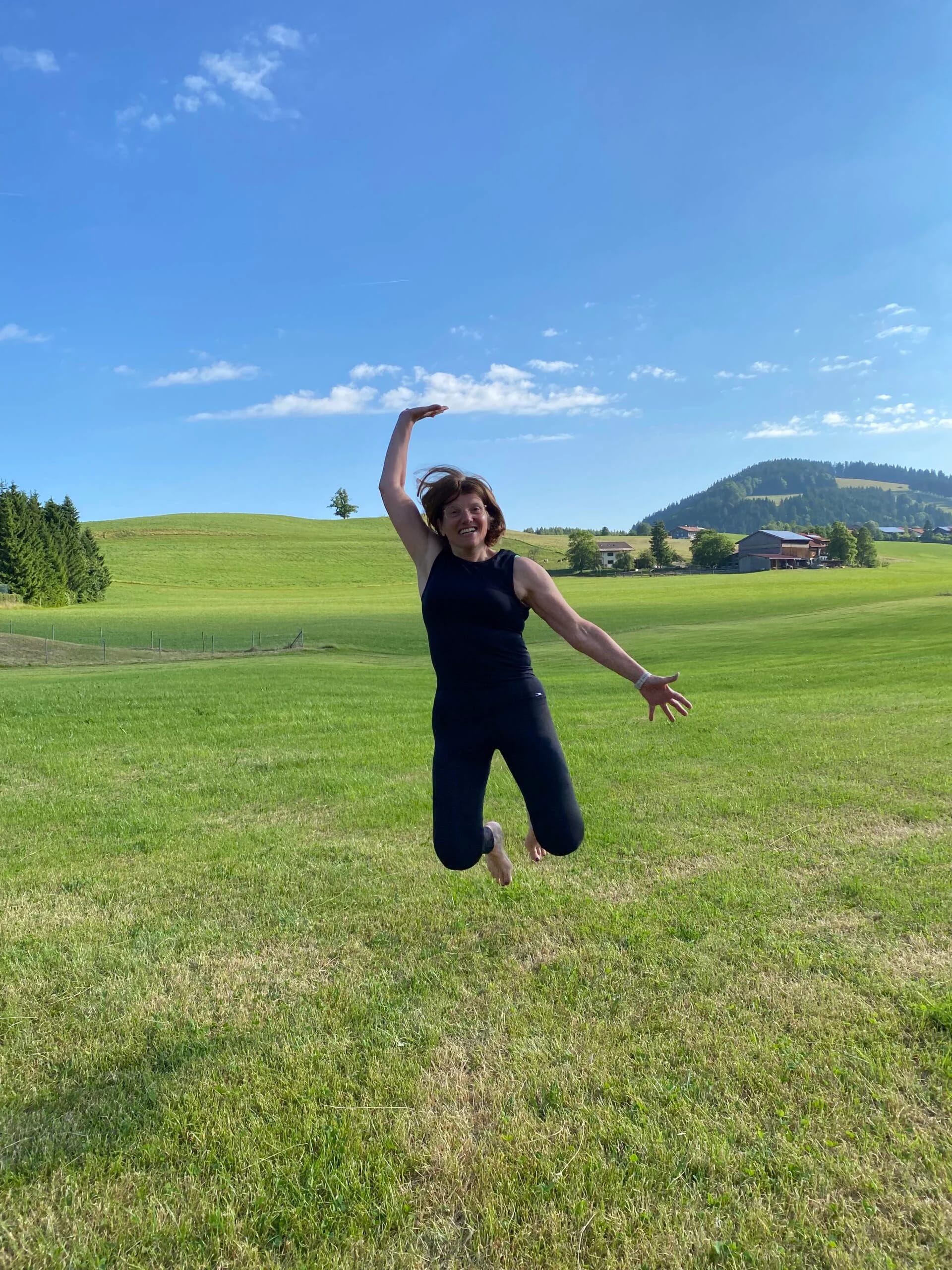 Sabine Wiemann springt freudig über eine grüne Wiese vor Bergpanorama
