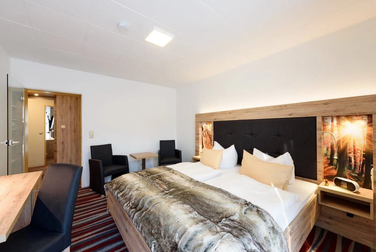 Hotelzimmer mit Doppelbett, Fellplaid, zwei Sesseln mit kleinem Tisch und Waldmotiv am Bettkopfende