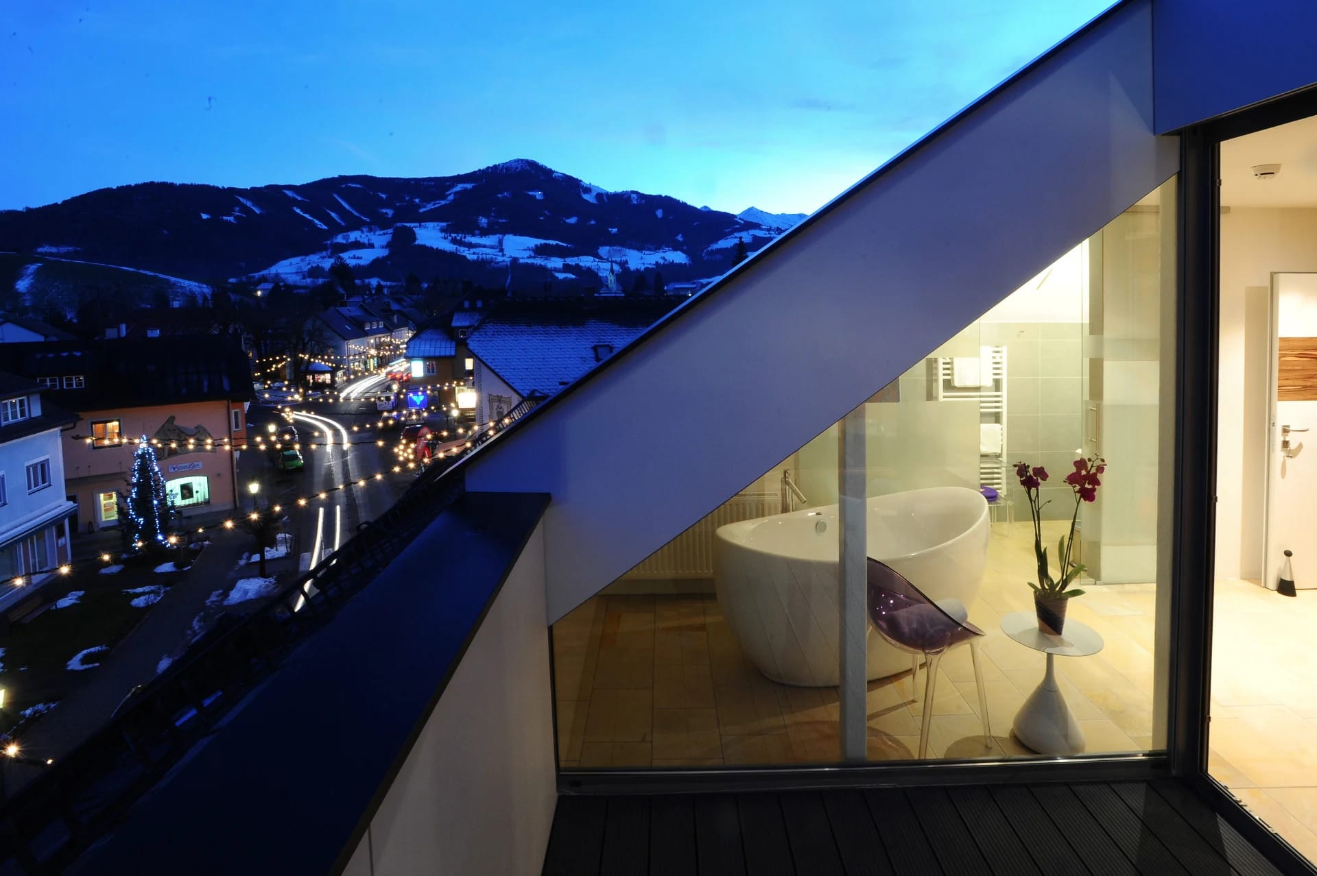 Loggia bei Nacht mit Blick auf den beleuchteten Ort und die Berge, dahinter verglastes Bad mit freistehender Wanne